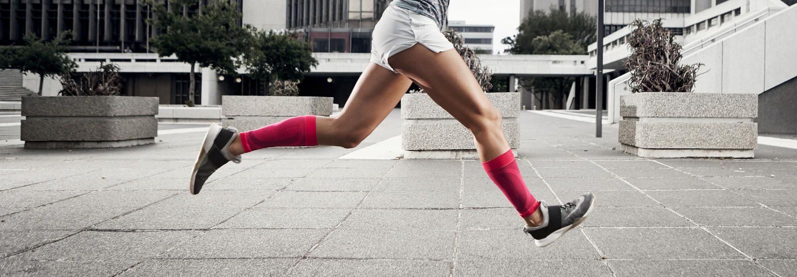 Breek door het grijs: onze compressie in mouwen in 7 kleuren Vrouw met roze kalveren loopt snel en met benen in de lucht door een stedelijke betonnen woestijn