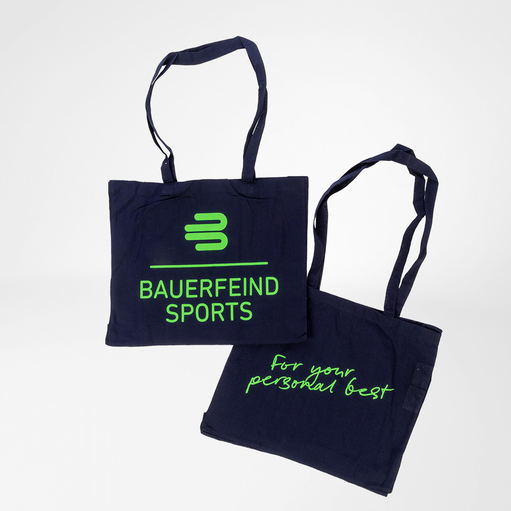 Twee donkerblauwe katoenen tassen van Bauerfeind Sports met felgroene opdruk. Eén toont het Bauerfeind Sports-logo, de andere de slogan „For your personal best“.