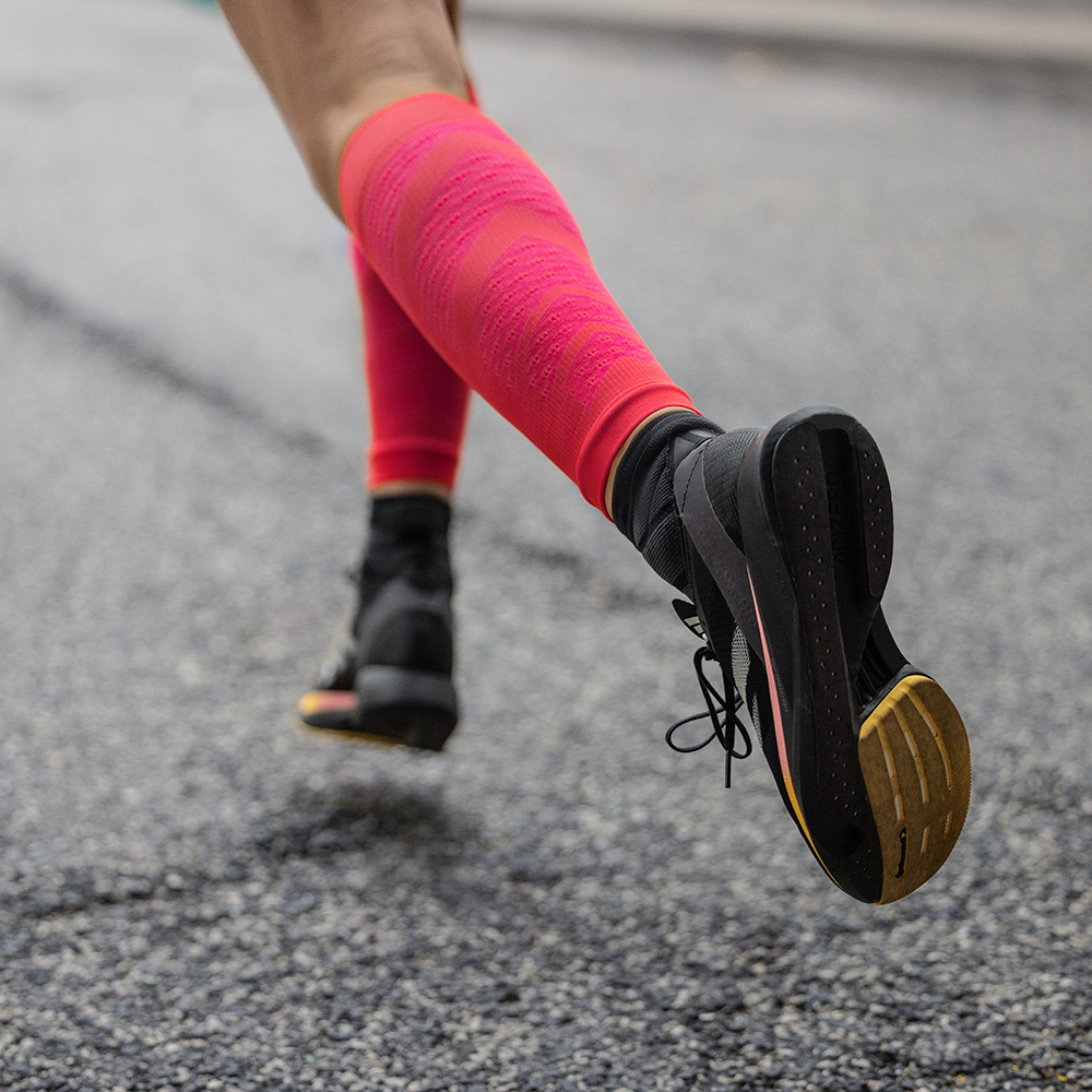 Close-up van een hardloopster die zich krachtig voortbeweegt op asfalt. Ze draagt zwarte hardloopschoenen en opvallende rode Run Performance Sleeves met compressie ter ondersteuning van de kuitspieren.