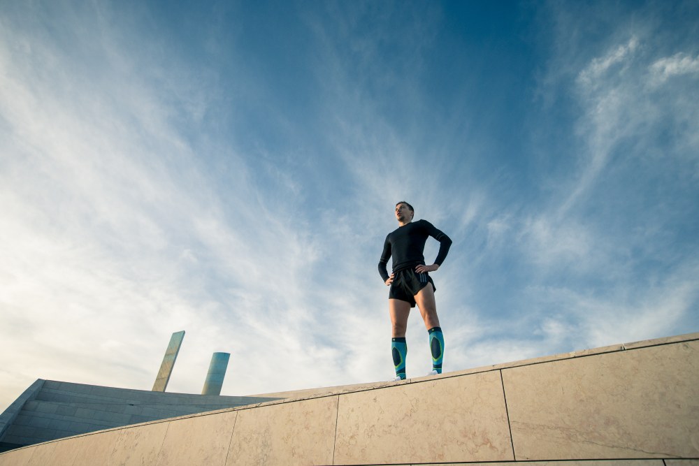 Runner met zwarte hardloopkleding en blauwloopsokken is bepaald op de muur van een modern gebouw en kijkt de verte in