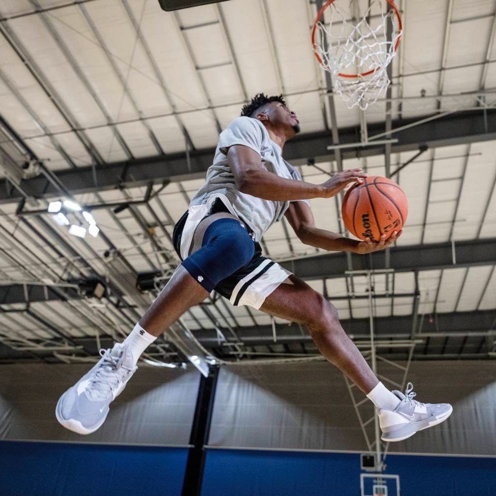 Vliegen met volledige ondersteuning – elke sprong telt! Een basketballer zweeft door de lucht voor een dunk, met een blauwe kniesteun om zijn rechterbeen.