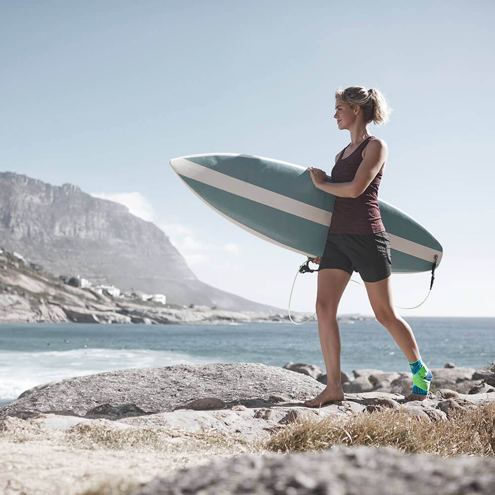 Vrouw met een surfplank onder haar arm met een enkelverband op het linkerbeen op een steenachtig strand met zee en bergen op de achtergrond