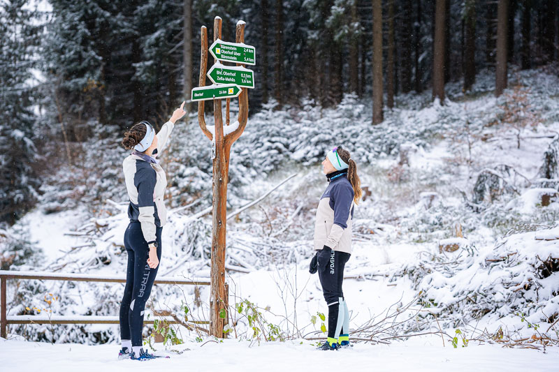 De juiste route vinden Twee vrouwen in winterhardloopkleding staan voor een houten wegwijzer in het besneeuwde bos. Een van hen wijst naar de bordjes terwijl de ander aandachtig kijkt. De wegwijzer geeft verschillende wandelroutes aan.