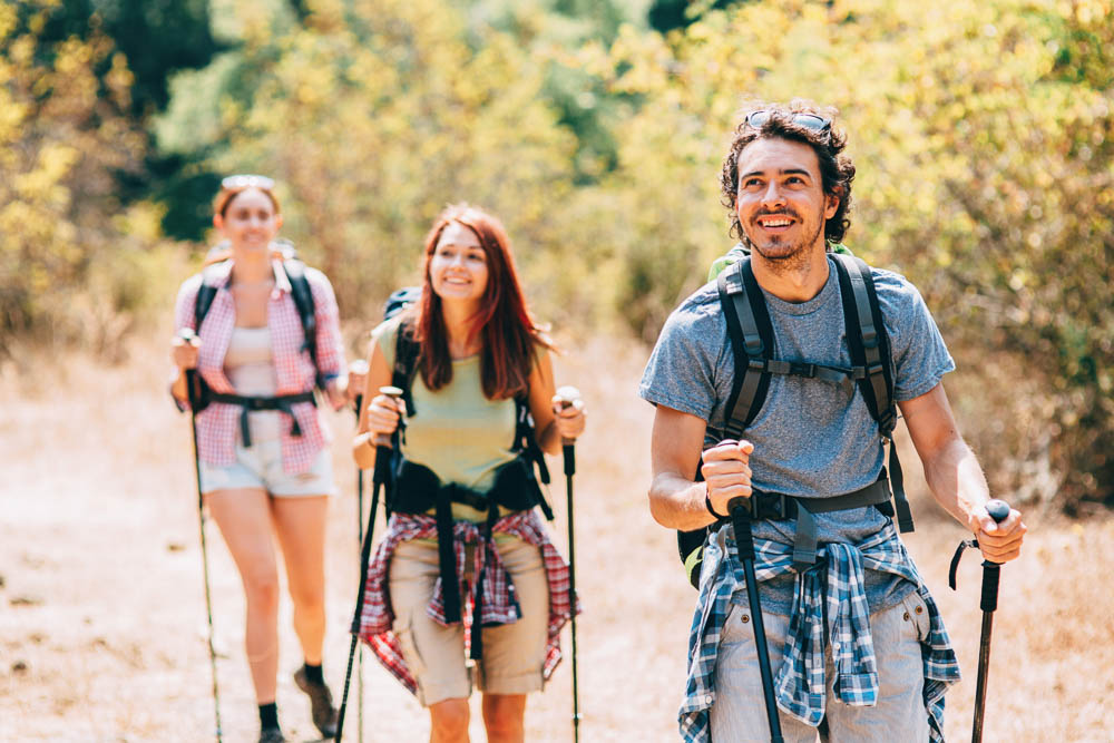 Wandelplezier op zonnig bospad Drie wandelaars, twee vrouwen en een man, lopen vrolijk met trekkingstokken over een zonnig bospad. Ze dragen sportieve kleding en rugzakken – de nadruk ligt op natuurlijke beweging en samen buiten zijn.