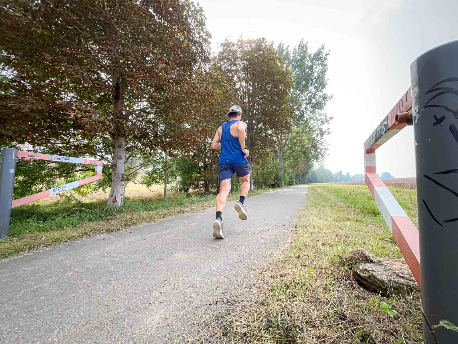 Hardlooptraining op Asfalt – Intensieve Trainingssessie in de Natuur. Een loper in een blauw shirt rent op een geasfalteerd pad in de natuur. Het pad wordt omgeven door bomen en een rood-witte barrière.