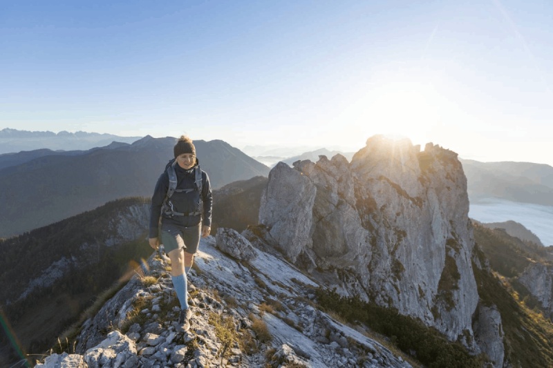 Licht wandelen - Outdoor Merino Compression Socks voor veeleisende tochten! Een wandelaar met Outdoor Merino Compression Socks loopt bij zonsopgang op een smal bergpad. Deze compressiesokken bieden optimale ondersteuning en comfort voor uitdagende wandelingen.