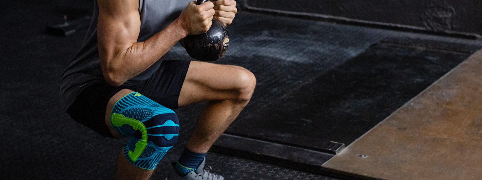 De sportknieondersteuning biedt u geweldige diensten, vooral in fitnesstraining Man met een kneebord gaat diep gehurkt met een kettlebell tussen de twee handen