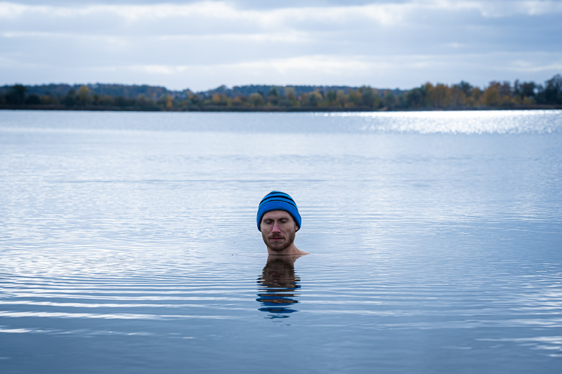 Man in meer tijdens ijsbad met blauwe muts Een man staat in een rustige meer, ondergedompeld tot aan zijn nek, met een blauwe muts op, en de herfstlandschap weerspiegelt in het water.