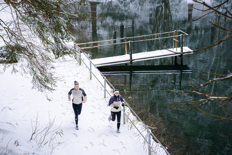 Winterrun bij het meer: Sporen in de sneeuw, reflecties in het water. Twee hardloopsters rennen langs een besneeuwd pad naast een meer met een met sneeuw bedekte steiger. Het wateroppervlak van het meer weerspiegelt de omliggende bomen. Beide hardloopsters dragen lichte winterloopjassen en zwarte broeken.