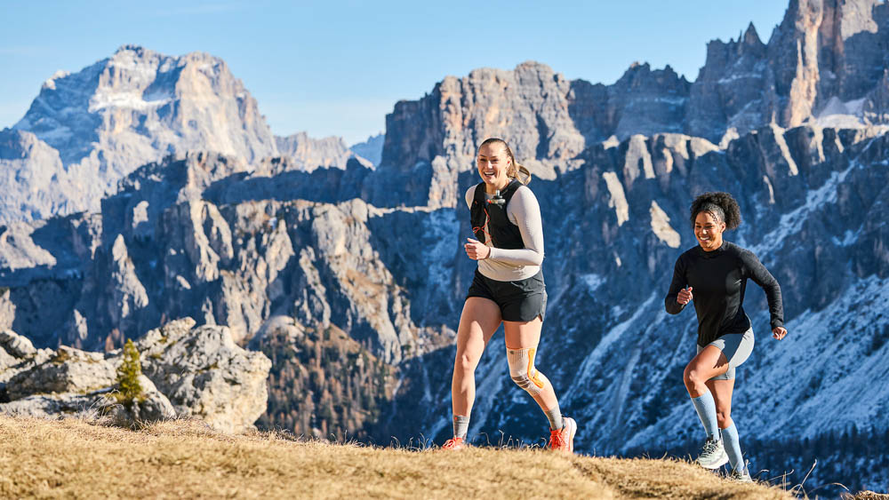 Twee loopsters trainen samen op een open bergrug met uitzicht op een indrukwekkend alpien landschap. Een van de atletes draagt een outdoor compressie-kniebandage. De foto toont beweging en sport in de natuur.
