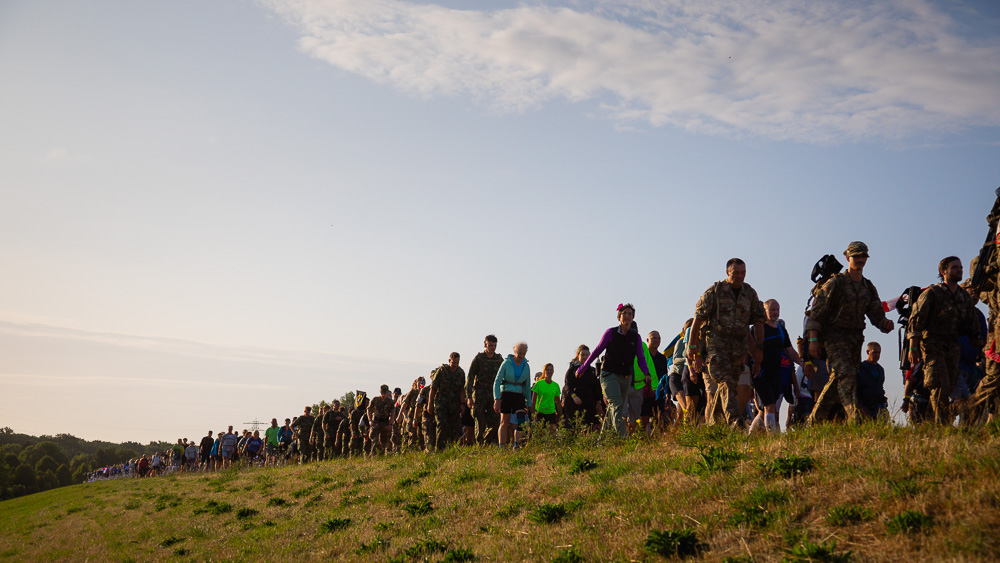 Wandelaars en soldaten in uniform lopen tijdens de 4Daagse in een lange rij over een dijk in het ochtendlicht.