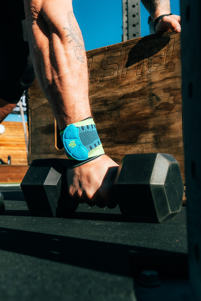 Close-up van een hand met een blauwe polsbandage die een zwarte dumbbell vastpakt; op de achtergrond staan een houten box en een outdoor rack in het zonlicht.