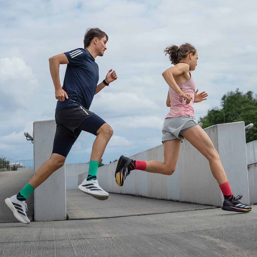 Twee hardlopers in beweging op een helling, een man in blauwe short met groene Bauerfeind-hardloopsokken en een vrouw in grijze short met rode Bauerfeind-hardloopsokken.