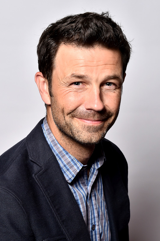 Dr. Casper Grim – Een vriendelijk portret met charisma Een man met kort, donker haar en een warme glimlach draagt een geruit overhemd en een donker colbert. Hij kijkt rechtstreeks in de camera tegen een neutrale achtergrond.