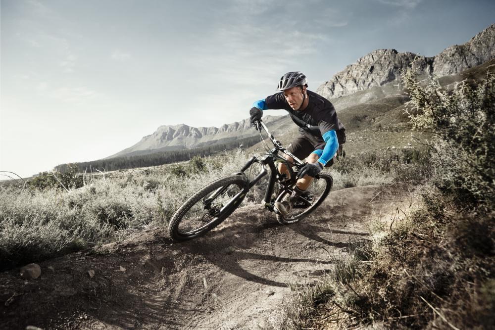 Man met Armenleeves rijdt op een mountainbike in een extreme hoek op een stoffig pad door een kaal landschap met struiken en bergen op de achtergrond