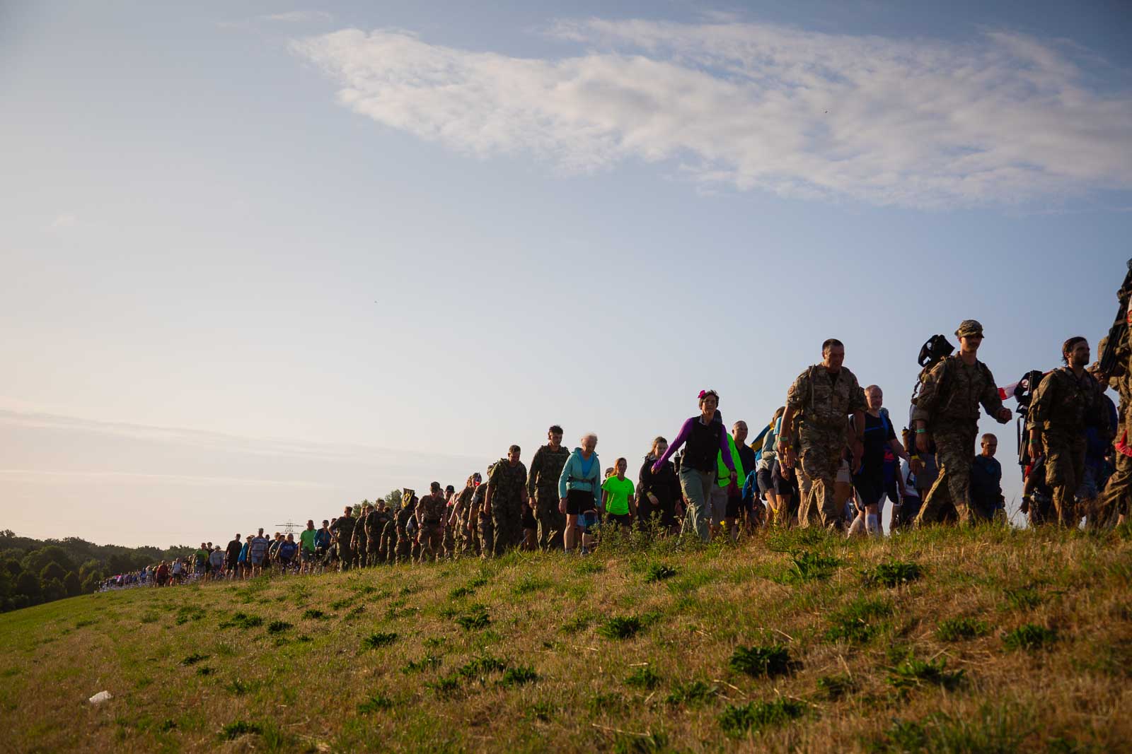Wandelaars en soldaten in uniform lopen tijdens de 4Daagse in een lange rij over een dijk in het ochtendlicht.