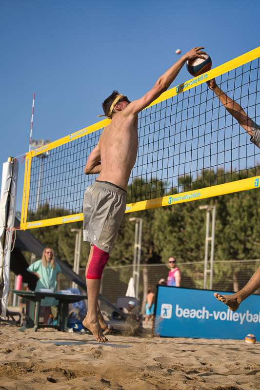 Een beachvolleybalspeler springt omhoog aan het net voor een blok, terwijl een tegenstander aan de andere kant de bal slaat. De speler draagt een felrode compressieband om zijn rechterbeen.