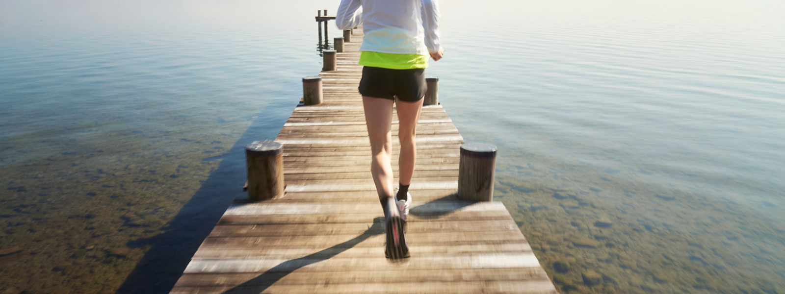 Pijn in de knie: waar kom je vandaan? Vrouw met een wit hardloopjasje en korte zwart lopende broek loopt over een voetgangersbrug naar het water