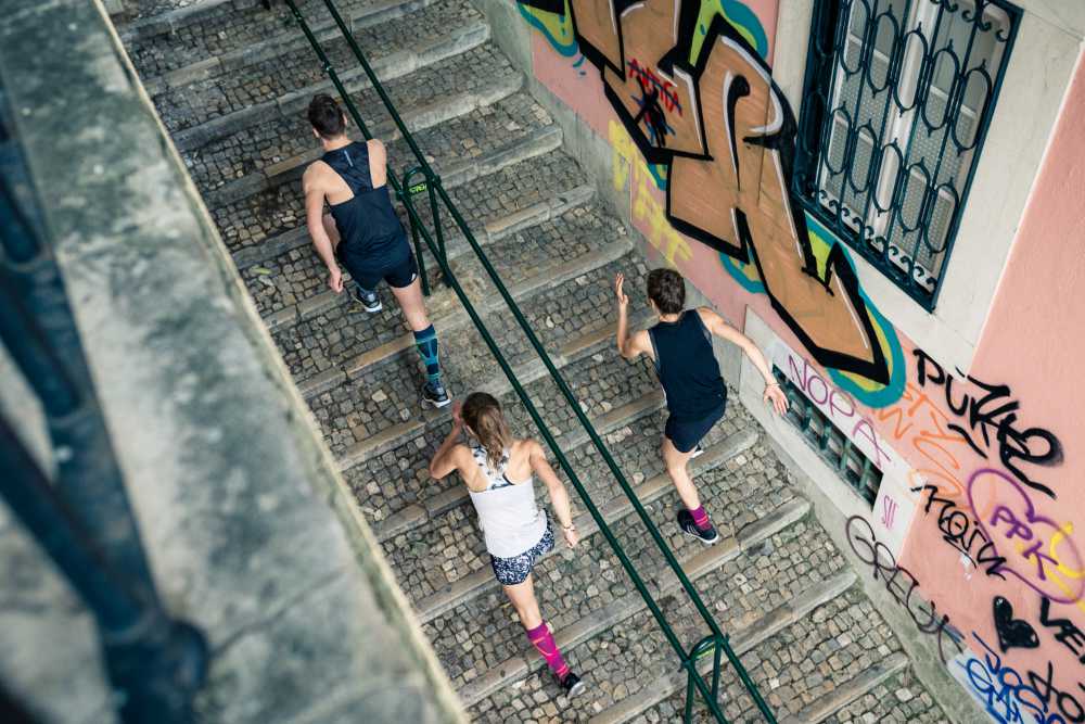 Bird's Eye View: een man en twee vrouwen in rennende outfits lopen een trap in een oude stad met graffiti op de muren