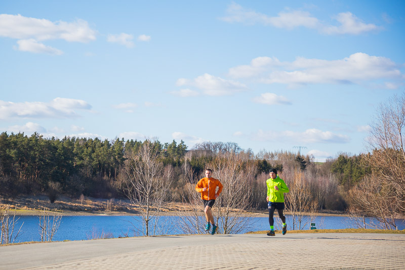 Frisse lucht en heldere gedachten – Hardlopen door de natuur Twee sporters rennen een zonnige heuvel op met uitzicht op een rustig meer en een bosrijke horizon. Beiden dragen felgekleurde hardloopjassen in oranje en geel.