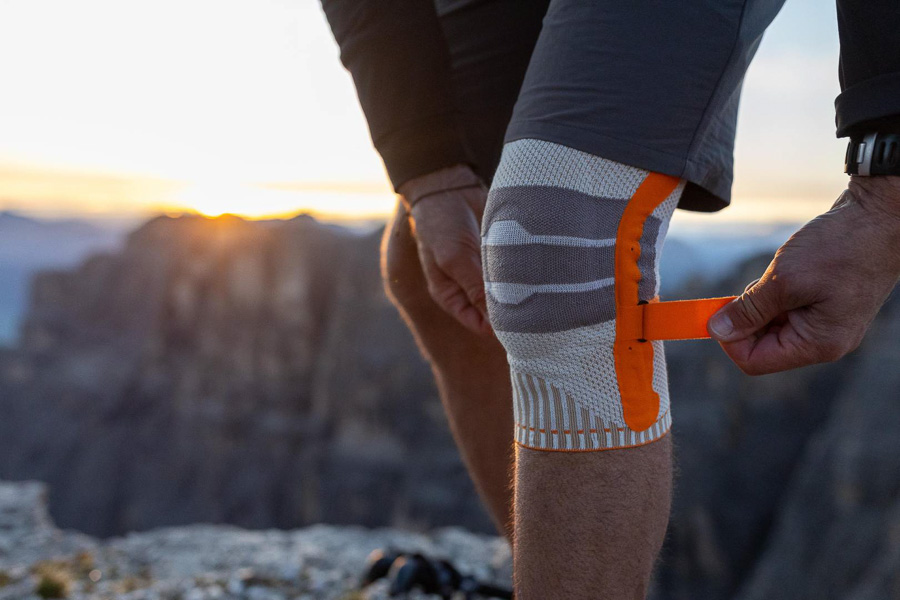 Optimale bescherming voor uitdagende wandelingen - Kniebrace voor stabiliteit en comfort! Een wandelaar past zijn oranje kniebrace aan tijdens een bergwandeling. Op de achtergrond zijn rotsachtige bergen en een zonsondergang te zien.