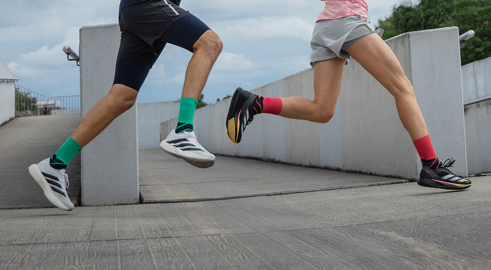 Vol energie vooruit Twee hardlopers in actie die een helling op sprinten. De man draagt groene compressiesokken met witte hardloopschoenen, de vrouw zwarte schoenen met rode compressiesokken. Beiden zijn vastgelegd midden in de beweging.