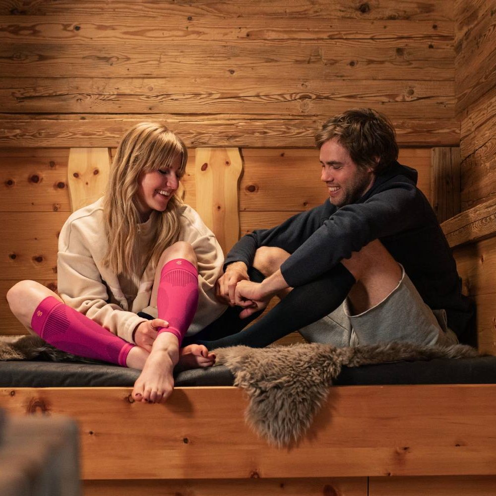 Ontspannen samen na een actieve dag Een glimlachend stel zit gezellig in een houten hut op een bank met een bontkleed. De vrouw draagt roze compressiekousen, terwijl de man zwarte draagt. Beiden genieten van hun welverdiende rust na een actieve dag.
