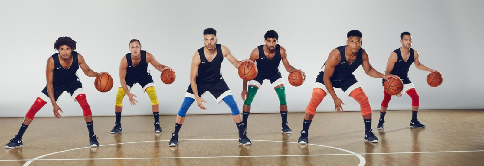Zes basketballers staan op een indoorveld in een rij en zijn gebukt in een voorbereidingshouding. Ze dragen zwarte shirts, witte shorts en compressiemouwen in verschillende kleuren om hun knieën.