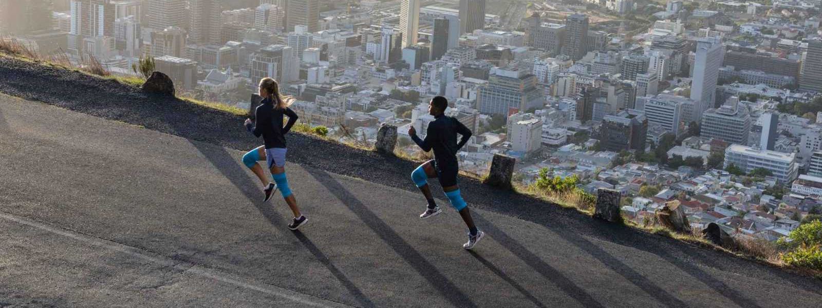 Zeer ontspannen boven de metropool dankzij de Bauerfeind sportcompressie knieondersteuning Een vrouw en een man met blauwe kniebars rennen een straat boven een grote stad