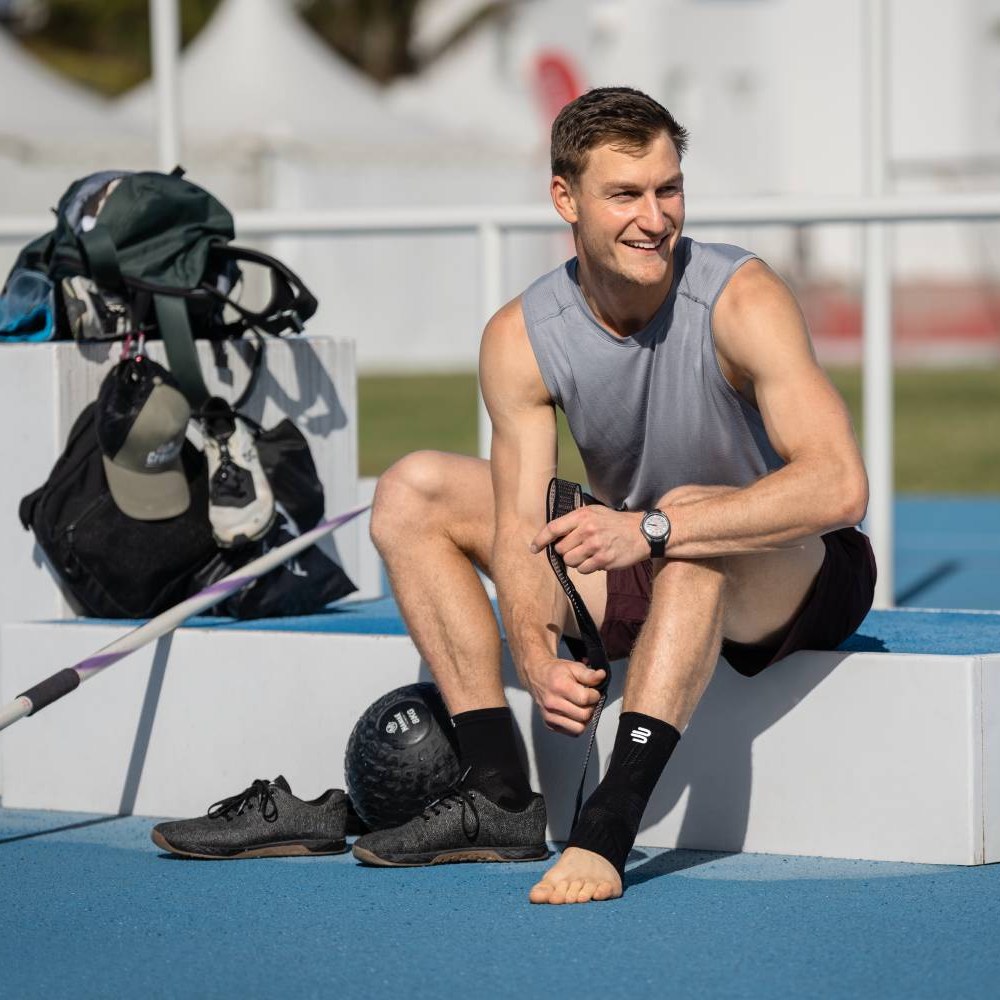 Klaar voor de volgende sprong – met stabiele enkels! Een atleet zit op een podium bij een atletiekbaan en trekt een Bauerfeind Sports Ankle Support brace over zijn enkel. Zijn sportschoenen en een trainingsbal liggen naast hem.