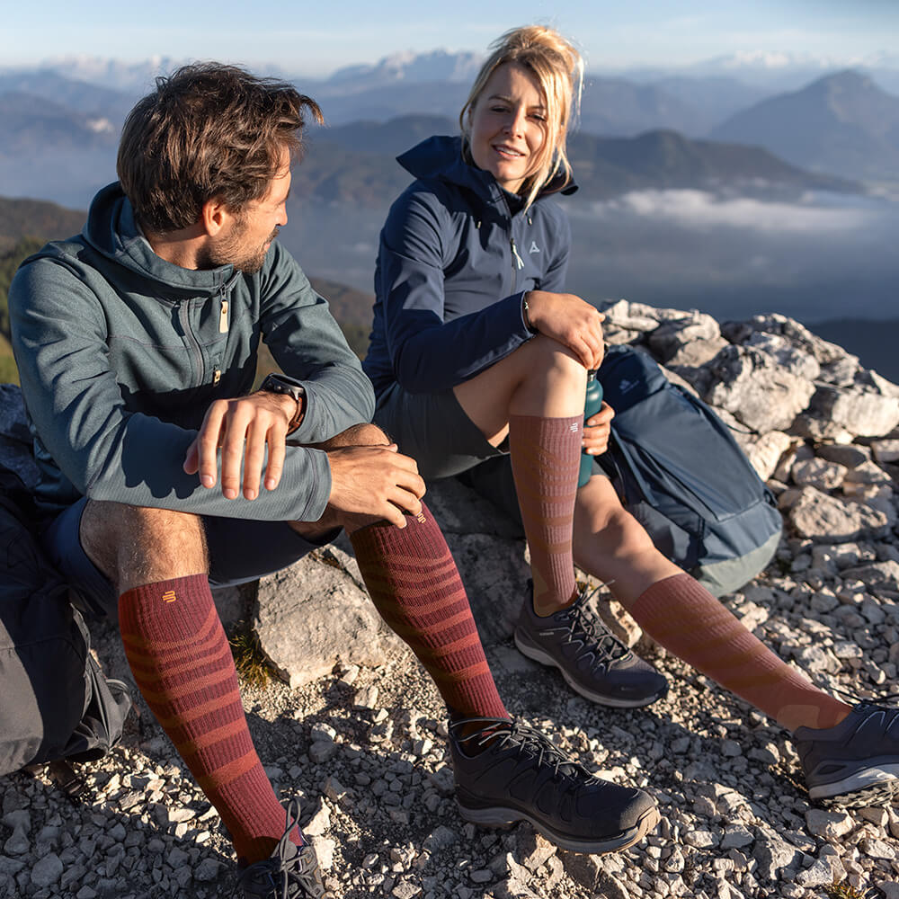 Twee wandelaars genieten van het uitzicht op een bergtop, terwijl ze Merino Outdoor compressiesokken in rood-bruin dragen. De sokken bieden optimaal comfort en ondersteuning voor lange tochten in uitdagend terrein.