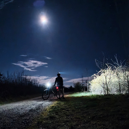 Avontuur bij maanlicht – als de uitdaging roept, houdt de nacht je niet tegen! Een fietser staat 's nachts op een bospad, verlicht door het volle maanlicht en een fietslamp. De scène toont de schaduw van de fietser, omringd door donkere silhouetten van bomen.