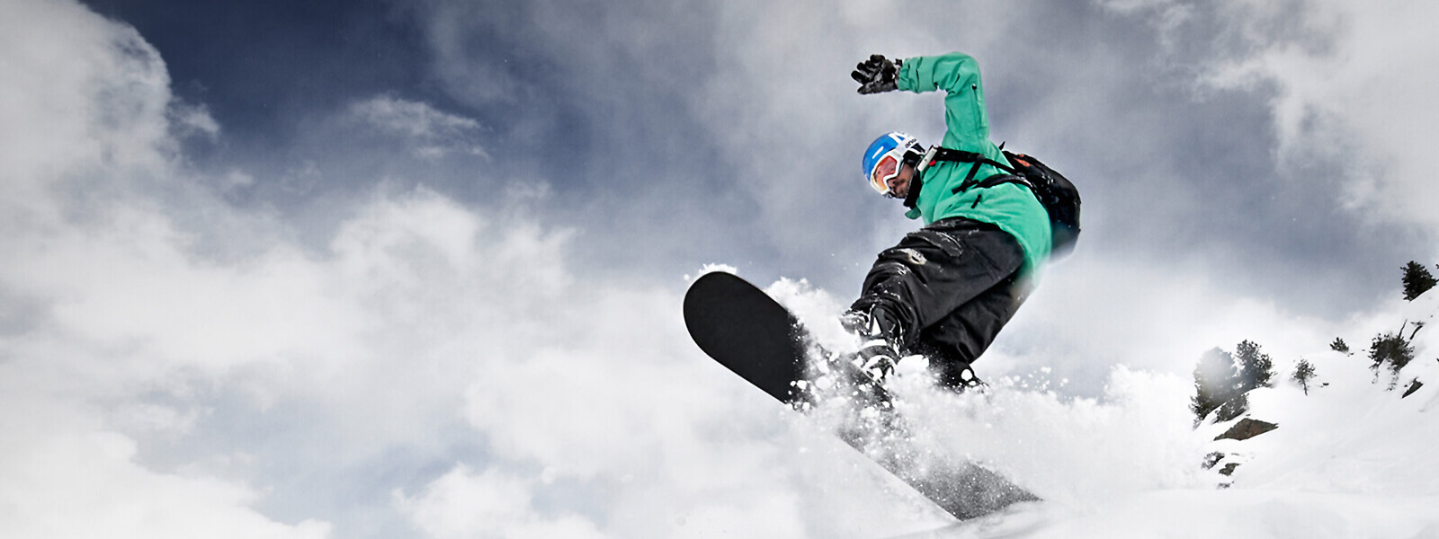 Kikkerperspectief: snowboarders in een groene jas komen van boven boven een kuif bord en veel gefluisterde sneeuw is te zien