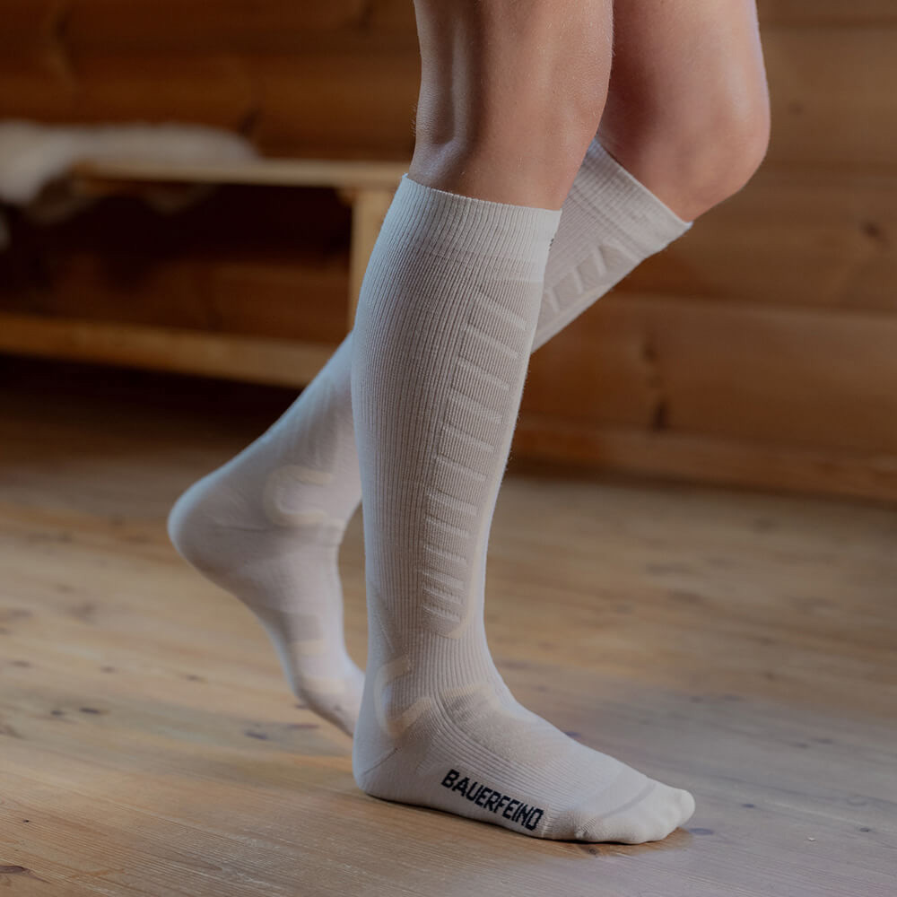 Perfecte pasvorm: Witte compressie-skisokken voor maximaal comfort op de piste Close-up van witte compressie-skisokken die tot onder de knieën komen. De persoon staat op een houten vloer in een hut. De sokken bieden extra comfort en ondersteuning met gewatteerde zones voor het skiën.