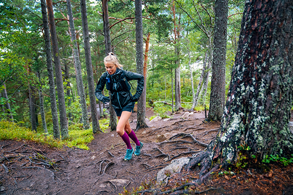 Traill Runner met langlopende sokken en trainingsjack loopt een bosweg op met veel wortels