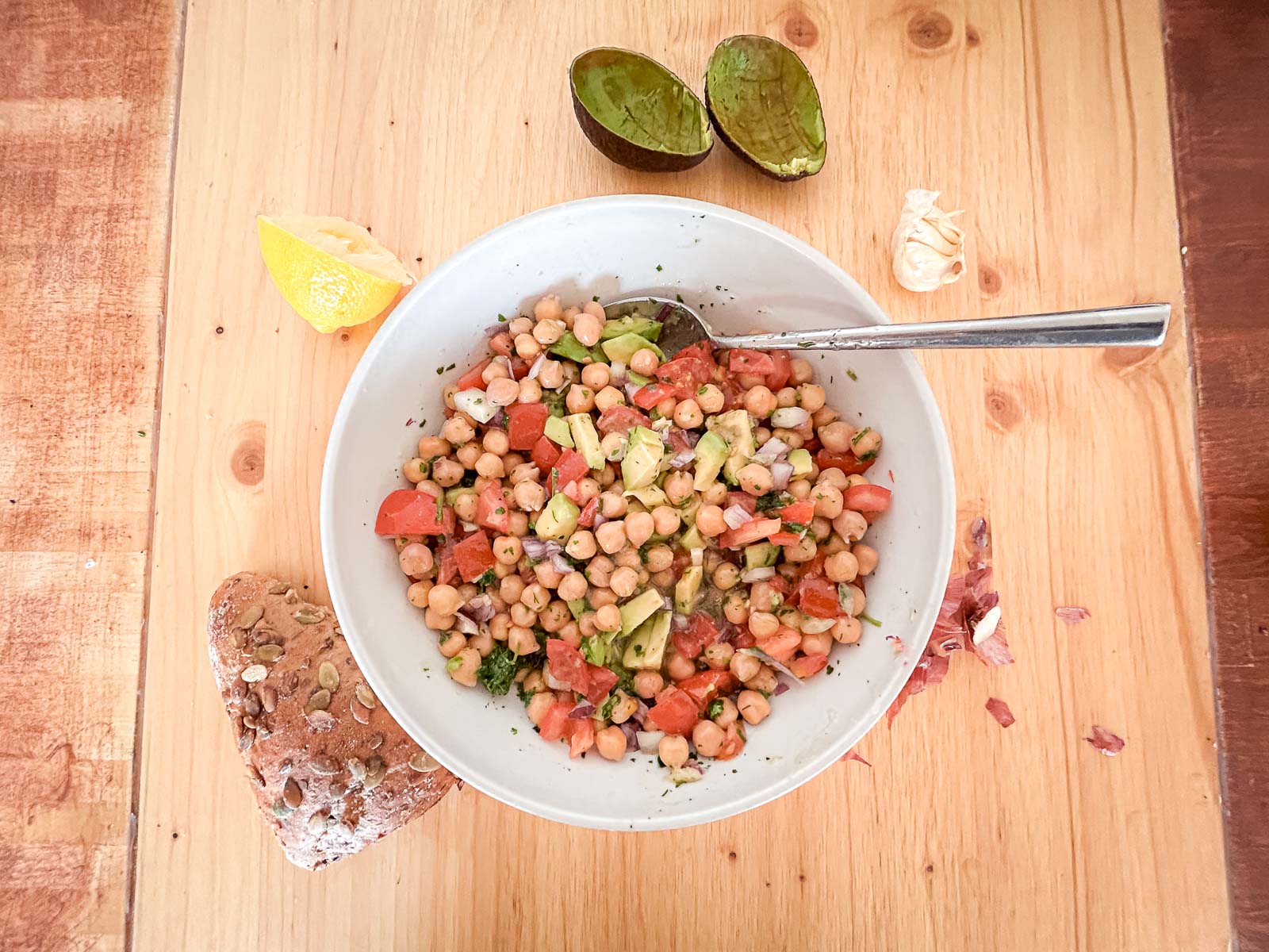 Kikkererwtensalade – Gezonde en Voedzame Maaltijd na de Training. Een kom kikkererwtensalade omgeven door verse ingrediënten zoals avocado, citroen en brood. Ideaal voor een gezonde en voedzame maaltijd na de training.