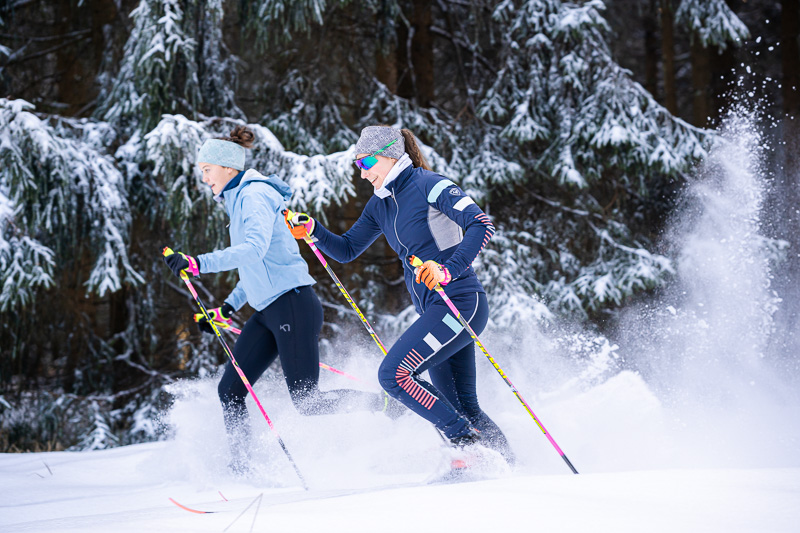 Dynamisch langlaufen in besneeuwd bos Twee vrouwen tijdens het langlaufen door met sneeuw bedekte bossen. Ze dragen kleurrijke wintersportkleding en glijden dynamisch door de sneeuw. De scène wordt gekenmerkt door dicht met sneeuw bedekte sparren, die het winterwonderland benadrukken.