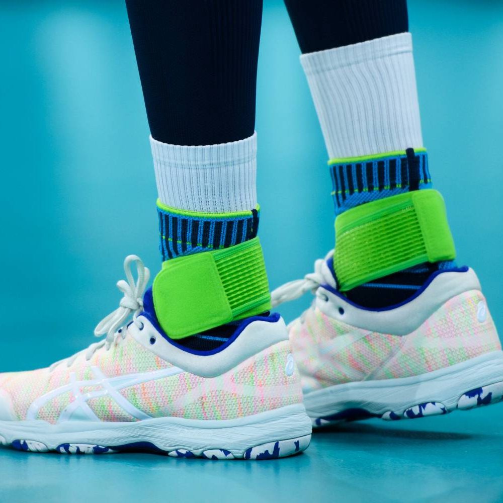 Springkracht met zekerheid – optimale ondersteuning voor intensieve wedstrijden! Close-up van de benen van een volleyballer op de indoorvloer. De Bauerfeind Sports Ankle Support Rivera wordt gedragen over hoge witte sokken.