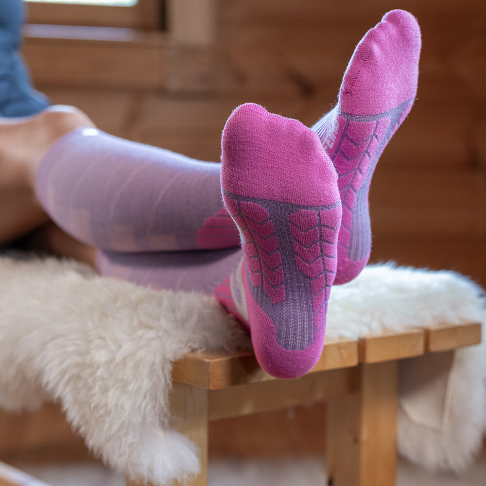 Een persoon ontspant op een bankje en draagt lila compressie-skisokken met roze zolen. De voeten zijn naar voren gestrekt, waarbij het gewatteerde zoolpatroon zichtbaar is.