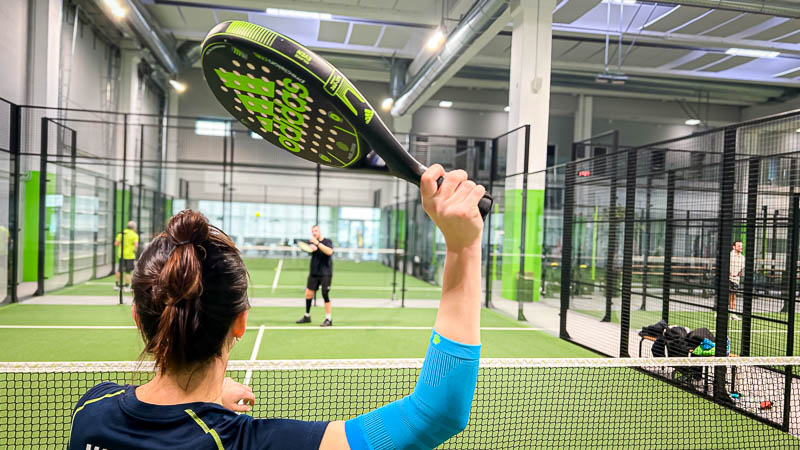 Een padelspeelster staat bij het net en maakt zich klaar om de bal te slaan. Haar arm is beschermd met een lichtblauwe compressiemouw.