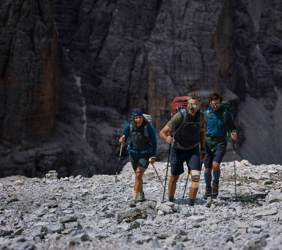 Samen uitdagend terrein trotseren - Wandelen met de juiste uitrusting! Een groep van drie wandelaars, uitgerust met rugzakken en trekkingstokken, beklimt rotsachtig terrein in de bergen. Een van de wandelaars draagt een kniebrace. Op de achtergrond zijn steile rotswanden te zien.