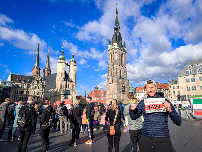 Een deelnemer van de Mitteldeutscher Halbmarathon houdt zijn startnummer omhoog op de marktplaats van Halle (Saale). Achter hem zijn de historische Rode Toren en Marktkirche te zien. Op de voorgrond bereiden lopers en toeschouwers zich voor op de race.