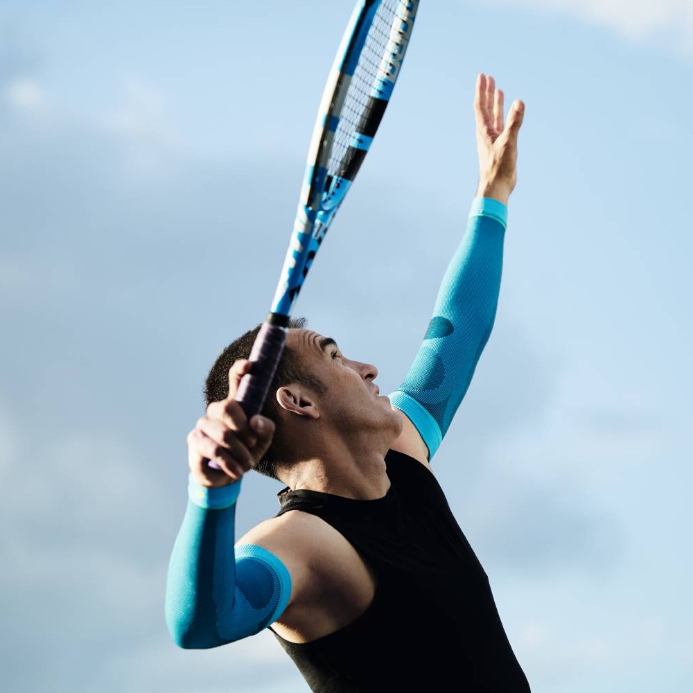 Tennisser met blauwe compressiemouwen bij de opslag Een tennisser met blauwe compressiemouwen serveert. De lucht is blauw met enkele wolken.