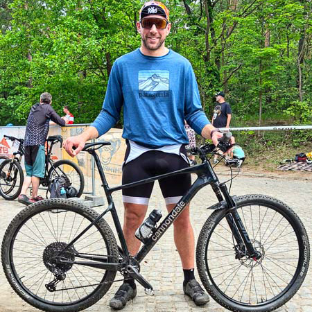 Gravel-glimlach: Na de uitdaging komt er altijd weer één! Een glimlachende man staat met een zwarte Cannondale-gravelbike op een bospad. Hij draagt een blauwe Patagonia trui met lange mouwen en een zwarte fietsbroek. Andere fietsers en groene bomen zijn op de achtergrond zichtbaar.
