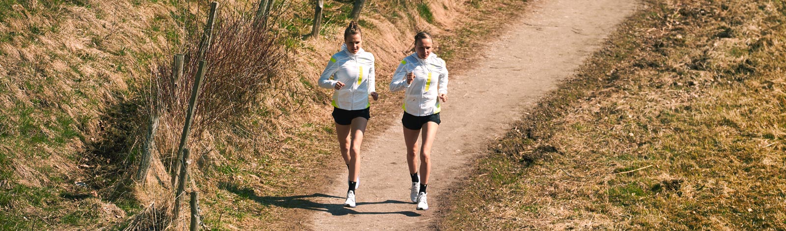 Twee lopers in heldere hardloopjassen lopen op een smal pad tussen spaarzaam overwoekerde weiden in de natuur