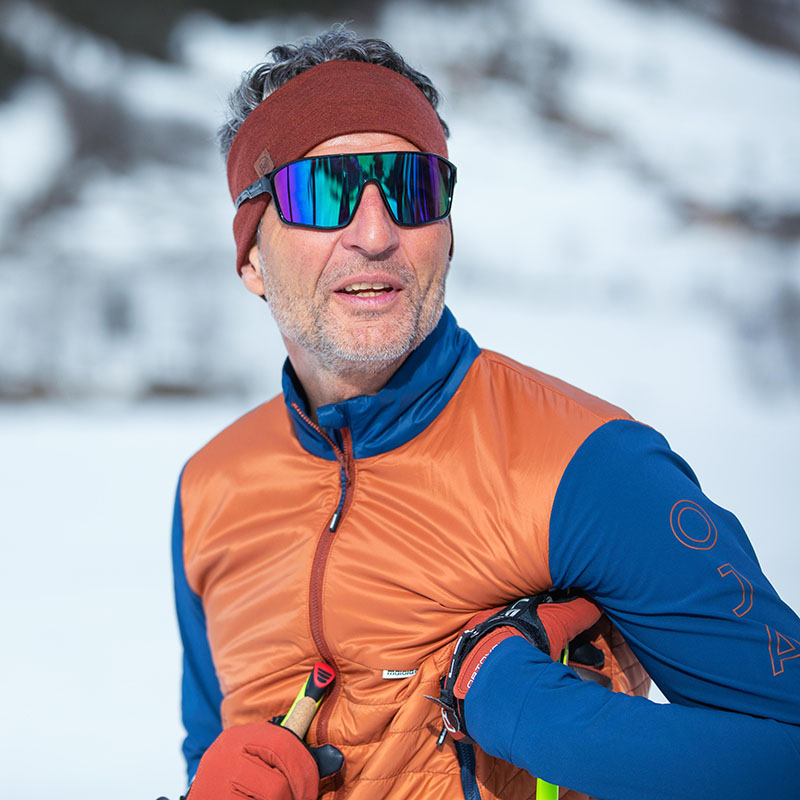 Portret van een skiër in het winterse bos Een oudere man met een skibril, een bruine hoofdband en een oranje-blauwe winterjas werd gefotografeerd in een portret voor een wit bos.
