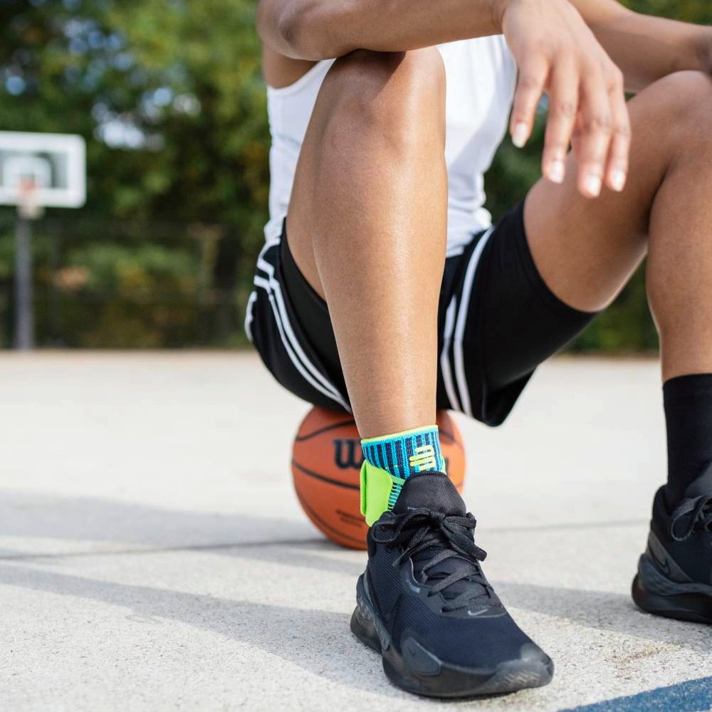 Stabiel op het veld – met stevige ondersteuning! Een basketballer zit op het veld met een basketbal onder zijn been. Hij draagt de Bauerfeind Sports Ankle Support Rivera in blauw-groen om zijn enkel.