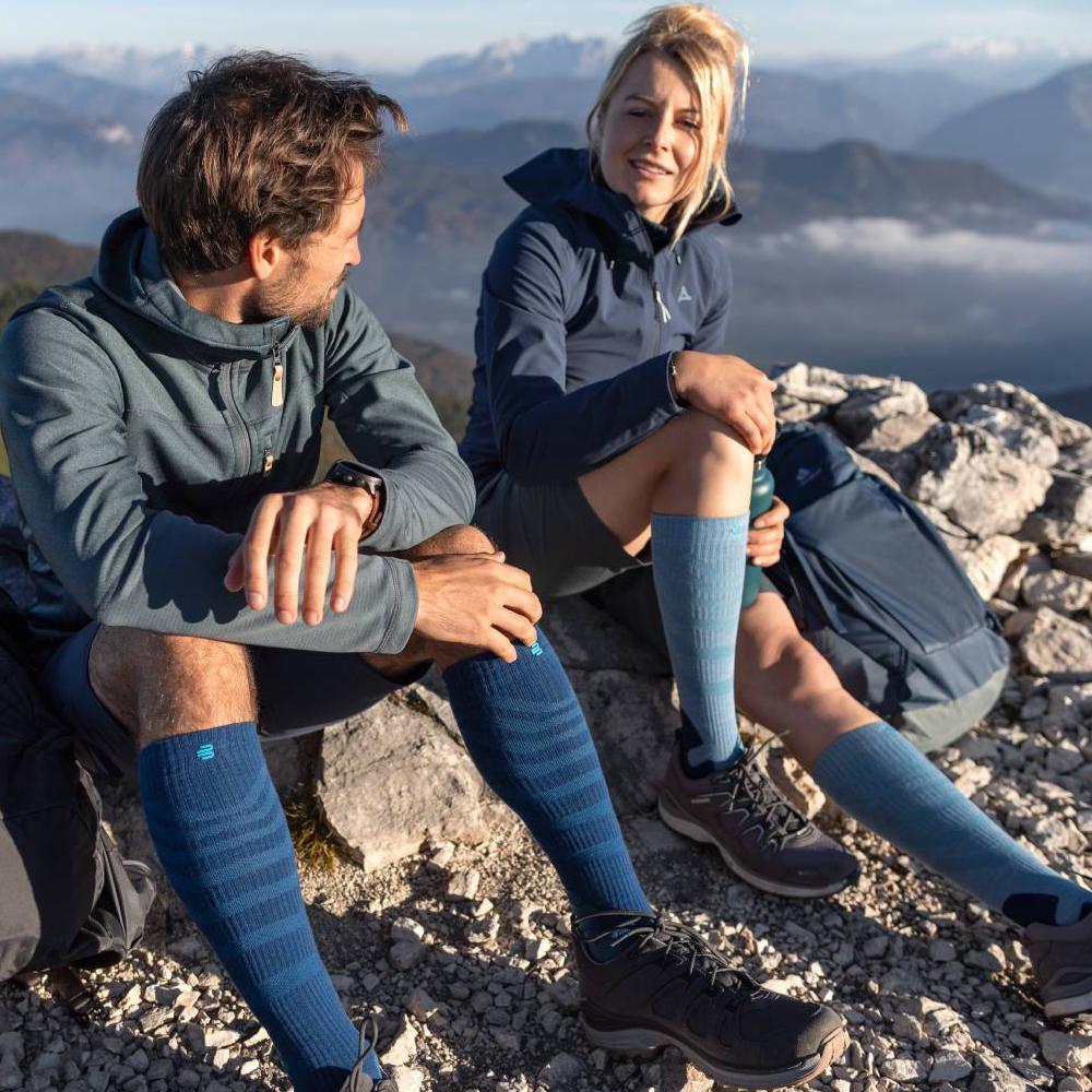 Hogerop met comfort – ontspanning op elke top. Twee wandelaars genieten van het uitzicht op een bergtop, terwijl ze Merino Outdoor compressiesokken in rood-bruin dragen. De sokken bieden optimaal comfort en ondersteuning voor lange tochten in uitdagend terrein.