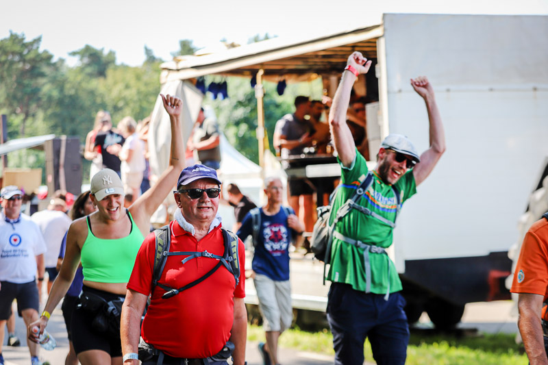Vrolijke deelnemers vieren feest tijdens de 4Daagse voor een muziekpodium. Een vrouw in een groen topje en een man in een rood shirt lopen lachend voorbij.