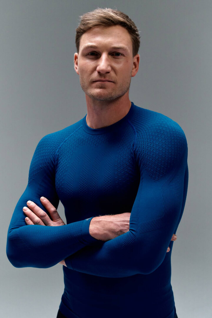 Een man in een nauwsluitend blauw sportshirt met gekruiste armen tegen een grijze achtergrond. De stof benadrukt de spieren en straalt kracht, controle en sportieve ambitie uit.